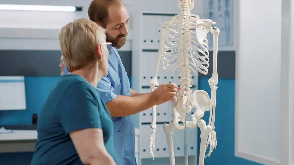 Médico hablando con la paciente y explicando el avance de la Osteoporosis, mediante un esqueleto anatómico.